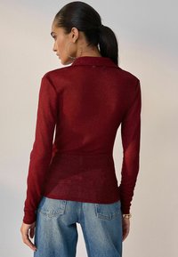 Rotes Langarmhemd mit Kragen, strukturiertem Stoff und figurbetontem Design. Getragen mit hellblauen hoch taillierten Jeans, dazu Ohrstecker mit Ohrhänger.