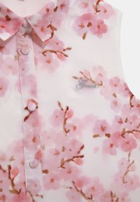 Blusa sin mangas con estampado de flores de cerezo en rosa, botones cubiertos de tela y pequeño logo bordado en plata en la parte superior del pecho.
