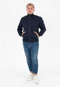 Marine blauwe zip-up sweatshirt met hoge kraag en voorkzakken, gecombineerd met lichtblauwe jeans en witte sneakers. Eenvoudig ontwerp en textiele stof.