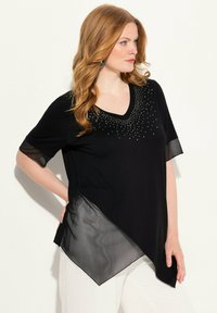 Top noir à col en V avec des manches courtes transparentes, présentant un design diagonal et des embellissements de petites clous sur le col avant et la zone de la poitrine.