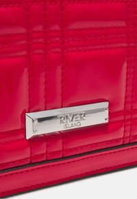 Rød, blank, quiltet materiale med et sølvmetal mærke prægede med "RIVER ISLAND" på en taske.