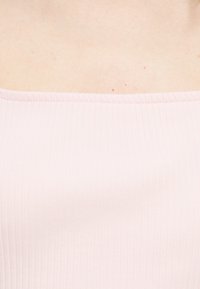 Κοντινή λήψη του πάνω μέρους του στήθους και των ώμων που φοράει ένα ανοιχτό ροζ ριμπ ζακέτα χωρίς ώμους, εμφανίζοντας φυσικό δέρμα με μερικές μικρές φακίδες.