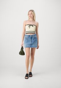Häkel-Bandeau-Top in Creme mit schwarzem Saum, kombiniert mit einem Jeans-Minirock mit fransigem Saum. Accessoires umfassen eine grüne Handtasche.