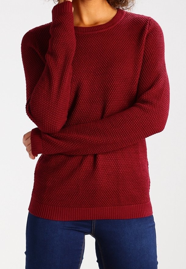 Femme portant un pull à manches longues texturé rouge foncé et un jean bleu foncé, avec un bras croisé et une main touchant son visage.