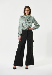Blusa di seta blu chiaro con motivi floreali e un fiocco al collo, abbinata a pantaloni cargo neri a vita alta con grandi tasche.