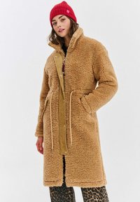 Žena v dlouhém hnědém kabátu, s červenou pletenou čepicí a kalhotami s leopardím vzorem, stojí s jednou rukou v kapse kabátu na bílém pozadí.