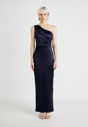 Robe de soirée en satin marine avec drapé sur une épaule, silhouette ajustée et texture subtile. Sandales métalliques complètent le look.