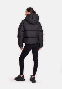 Giacca a vento nera con cappuccio, caratterizzata da una texture trapuntata e un design accorciato. Modella abbinata a leggings neri e sneaker chunky.