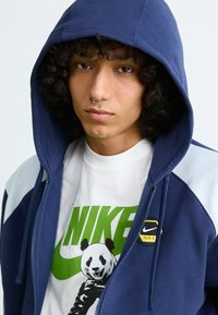 Navy blue zip-up hoodie with gray accents, featuring a logo patch. Underneath is a white t-shirt with a green Nike graphic and panda design.