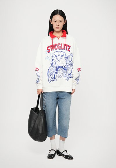 Felpa bianca con dettagli rossi, con una grafica di gufo e il testo "STEGLLITZ". Abbinata a pantaloni cropped in denim chiaro e una borsa nera.