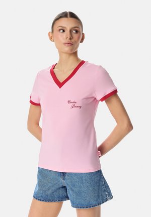 Noor naine kannab roosat V-kaelusega T-särki punase äärisega ja teksapükse, seisab käed selja taga, vaadates kõrvale.