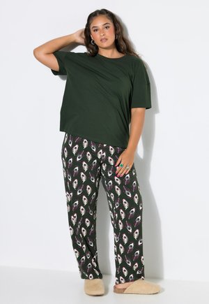 Joven mujer con camiseta oversize verde oscuro, pantalones con estampado de plumas de pavo real, zapatillas beige, de pie con una mano detrás de la cabeza.