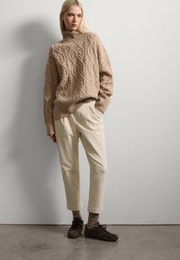 Suéter de punto beige con cuello alto, de corte holgado, combinado con pantalones crema de corte tapered y zapatillas marrón oscuro. Materiales suaves y texturizados.