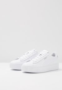 Baskets blanches avec une tige en cuir lisse, bout rond, design à lacets et semelle plate épaisse. Comprend des détails perforés et un marquage discret.