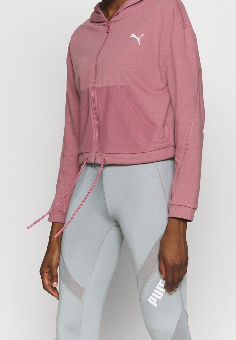 Personne portant un sweat à capuche zippé mauve avec logo Puma et des leggings Puma gris clair avec des panneaux en mesh sur fond blanc.