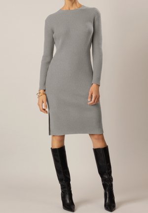 Femme portant une robe grise cintrée à côtes, longueur genou, manches longues, et des bottes en cuir noires montant jusqu'aux genoux, debout devant un fond uni.