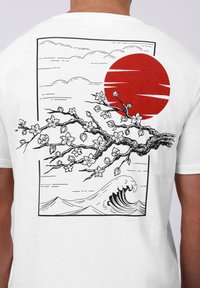 Λευκό μπλουζάκι με μαύρη και κόκκινη γραφική παράσταση άνθεων κερασιάς, κόκκινου ήλιου, κυμάτων και σύννεφων σε ορθογώνιο σχέδιο.