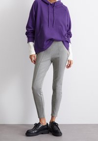 Felpa viola con tasca frontale, abbinata a pantaloni grigi a gamba stretta con cuciture laterali e scarpe nere stringate con suola spessa.