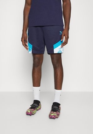 Homme portant un short de sport bleu marine avec des accents turquoise et blancs, des chaussettes blanches et des crampons noirs avec des motifs rose néon et vert.