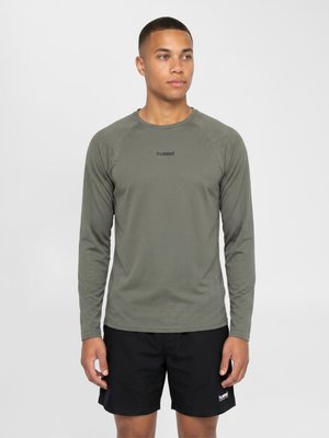 Junger Mann, der ein olivgrünes Langarmshirt und schwarze Shorts trägt, steht frontseitig vor einem schlichten hellen Hintergrund.