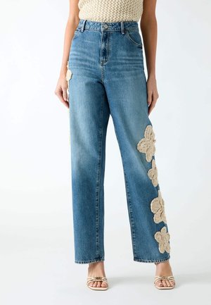 Vrouw draagt blauwe high-waisted spijkerbroek met beige gehaakte bloemenpleisters langs de buitenkant van de benen en beige open-toe sandalen.
