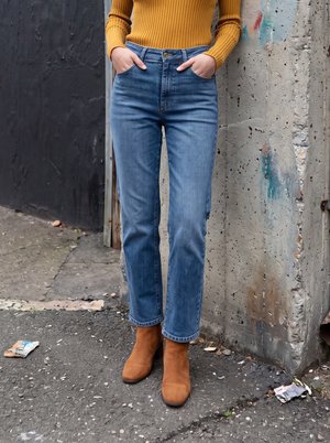 Person iført blå jeans, brune ankelstøvler og en sennepsfarvet ribbet sweater med hænderne i lommerne står på en revnet fortorv.