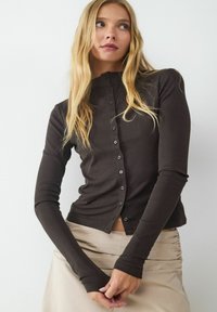 Braunfarbener Langarmoberteil mit hohem Kragen und durchgehender Knopfleiste, mit einer taillierten Passform und weichem, dehnbarem Material. Kombiniert mit einem beigen Rock.