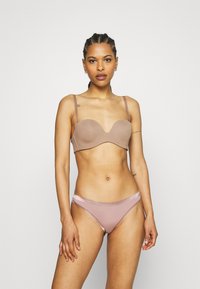 Bralette em tecido nude suave com um design de curvas subtil. Cueca combinando com uma faixa rosa clara e detalhes transparentes. Alças ajustáveis.