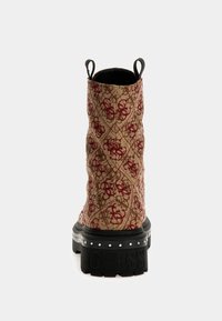 Une botte de cheville avec une tige en tissu beige ornée de détails en rouge, un design matelassé et une épaisse semelle en caoutchouc noir estampillée "GUESS."