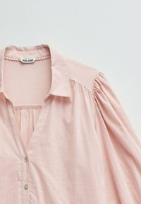 Blusa de manga larga color rosa claro con hombros fruncidos, cuello abierto y dos botones delanteros, con la etiqueta "Salsa Jeans" en el interior del escote.