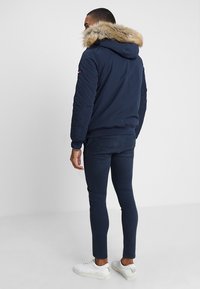 Man i marinblå jacka med pälsfodrad huva, marinblå smala byxor och vita sneakers, står mot en enfärgad vit bakgrund.