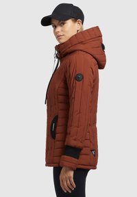 Hnedá puffa bunda s kapucňou, horizontálne prešité design, čierne akcenty, bočné vrecká a nášivka loga na rukáve. Nosí sa čierna čiapka.