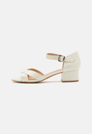 Sandal de couro creme com design de tiras cruzadas, fecho de fivela no tornozelo e um salto bloco largo. Textura suave e silhueta minimalista.