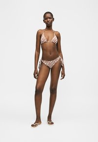 Femme debout portant un bikini marron et blanc à motifs avec des liens sur les côtés et des tongs blanches contre un fond blanc uni.