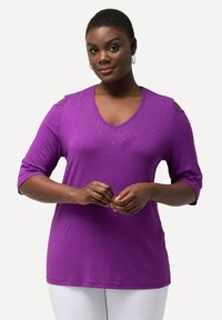 Paarse V-hals top met driekwart mouwen, voorzien van cold shoulder uitsparingen en decoratieve rhinestones langs de halslijn. Glad stofgevoel.