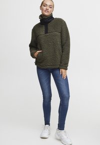 Olijfgroen fleece pullover met zwarte kraag en borstpaneel, lange mouwen, voorzak, gecombineerd met blauwe skinny jeans en witte sneakers.
