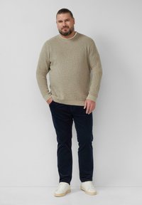 Beige geripptes Sweatshirt mit Rundhalsausschnitt, kombiniert mit dunkelblauen Hosen. Die Schuhe sind weiße Sneakers. Das Model steht mit den Händen in den Taschen.
