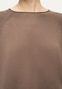 Gros plan sur un pull en tricot marron clair montrant l'encolure et le haut de la poitrine d'une personne.