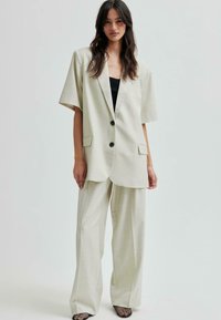 Blazer oversized beige chiaro con maniche corte, due bottoni e tasche frontali, abbinato a pantaloni palazzo dello stesso colore.