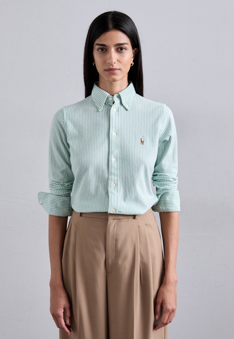 Camicia button-up a righe verde chiaro con colletto, con maniche arrotolate; abbinata a pantaloni beige a vita alta. Piccolo emblema sulla camicia.