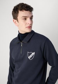 Hanorac din fleece bleu marin cu guler tip fermoar pe jumătate, având o pată albă în formă de scut cu text. Textură netedă, design casual.