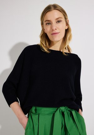 Frau mit blonden Haaren, die ein schwarzes langärmeliges Oberteil und einen hoch taillierten grünen Rock mit gebundenem Gürtel trägt, steht vor einem schlichten hellen Hintergrund.