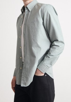 Mężczyzna w jasnozielonej koszuli na guziki na białym t-shircie i czarnych spodniach, z jedną ręką w kieszeni, na jednolitym białym tle.