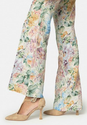 Pantalones anchos con estampado floral combinados con tacones altos de ante beige de tira en T con hebilla adornada con joyas, usados por una modelo que está de pie sobre un fondo blanco.