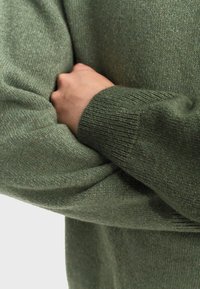 Maglione verde a maglia con una texture morbida, caratterizzato da polsini a coste e una vestibilità ampia. Dettaglio dell'avambraccio che attraversa il petto.