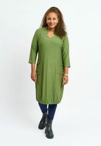 Grön knälång klänning med strukturerad mönster, V-hals, trekvartslånga ärmar och sidofickor, kombinerad med marinblå leggings och svarta stövlar.