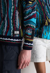 Colorful knitted sweater featuring intricate patterns of red, green, blue, and yellow, with ribbed cuffs and a logo patch on the lower sleeve.