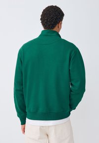 Grøn sweatshirt med høj krave, struktureret stof og ribstrikkede ærmekanter. Kombineres med en hvid lagdelt skjorte under, set bagfra.