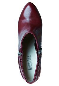 Zapato de cuero burdeos con punta afilada y paneles laterales de malla texturizada. Presenta un acento metálico cerca de la abertura.