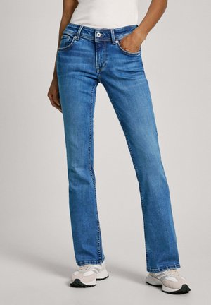 Person wearing blue straight-leg jeans with a white top and white sneakers, standing with one hand in pocket against a plain background.
